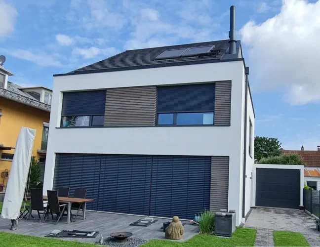 Modernes zweistöckiges Haus mit großen blauen Fensterläden, Sitzgelegenheiten im Freien und einem gepflegten Hof unter blauem Himmel.