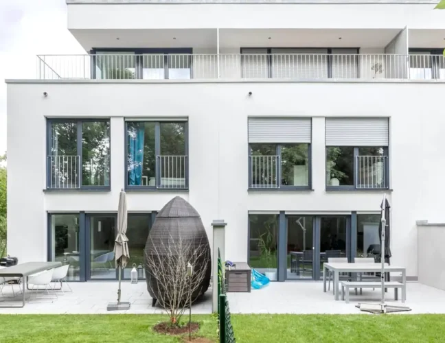 Modernes, weißes Apartmenthaus mit großen Fenstern und einem Innenhof mit Gartenmöbeln und grünem Rasen.