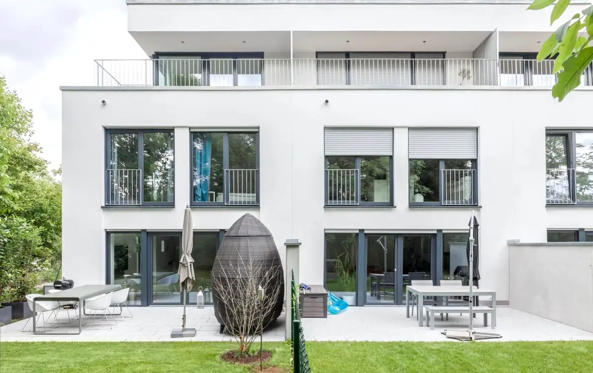 Modernes, weißes Apartmenthaus mit großen Fenstern und einem Innenhof mit Gartenmöbeln und grünem Rasen.