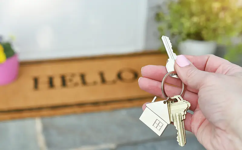 Eine Hand hält Hausschlüssel über einer "HELLO"-Fußmatte - Symbolbild für schlüsselfertige Massivhäuser.