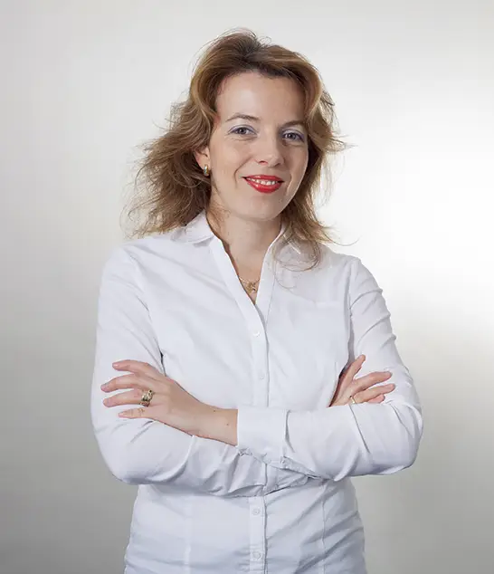 Ada Jac mit hellbraunem Haar im weißen Hemd, selbstbewusst lächelnd - perfekt für Ihr nächstes Massivhausprojekt.