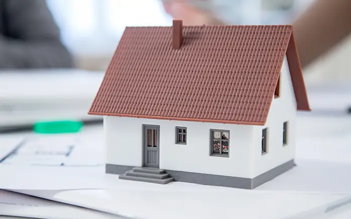 Ein kleines Modellhaus mit rotem Dach liegt auf einem unscharfen Bürotisch auf dem Papier.