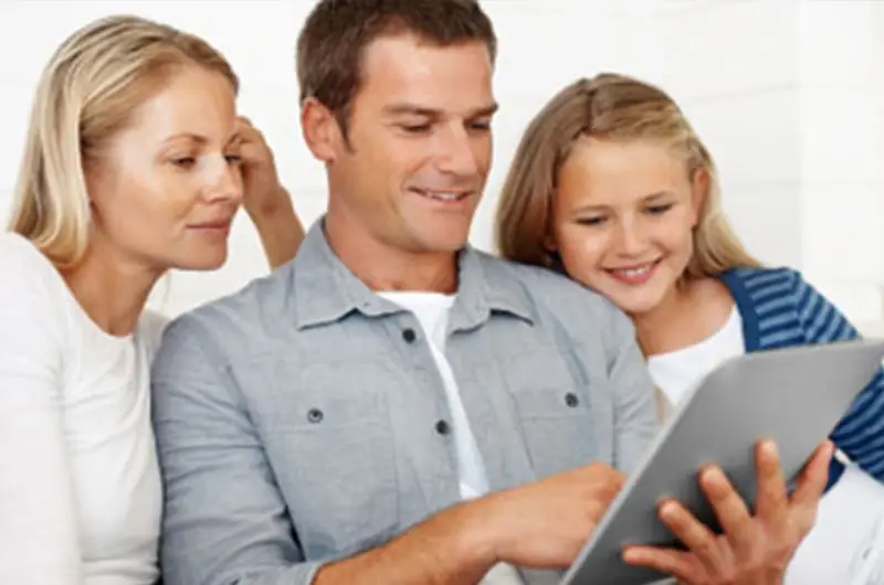 Eine lächelnde dreiköpfige Familie genießt die gemeinsame Zeit, während sie auf der Couch sitzend auf einem Tablet Ideen für energieeffizientes Bauen erforscht.